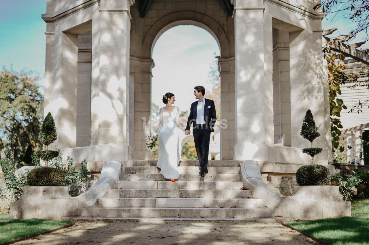 Un couple en tenue de mariage descend des marches en pierre devant une arche dans un jardin ensoleillé.