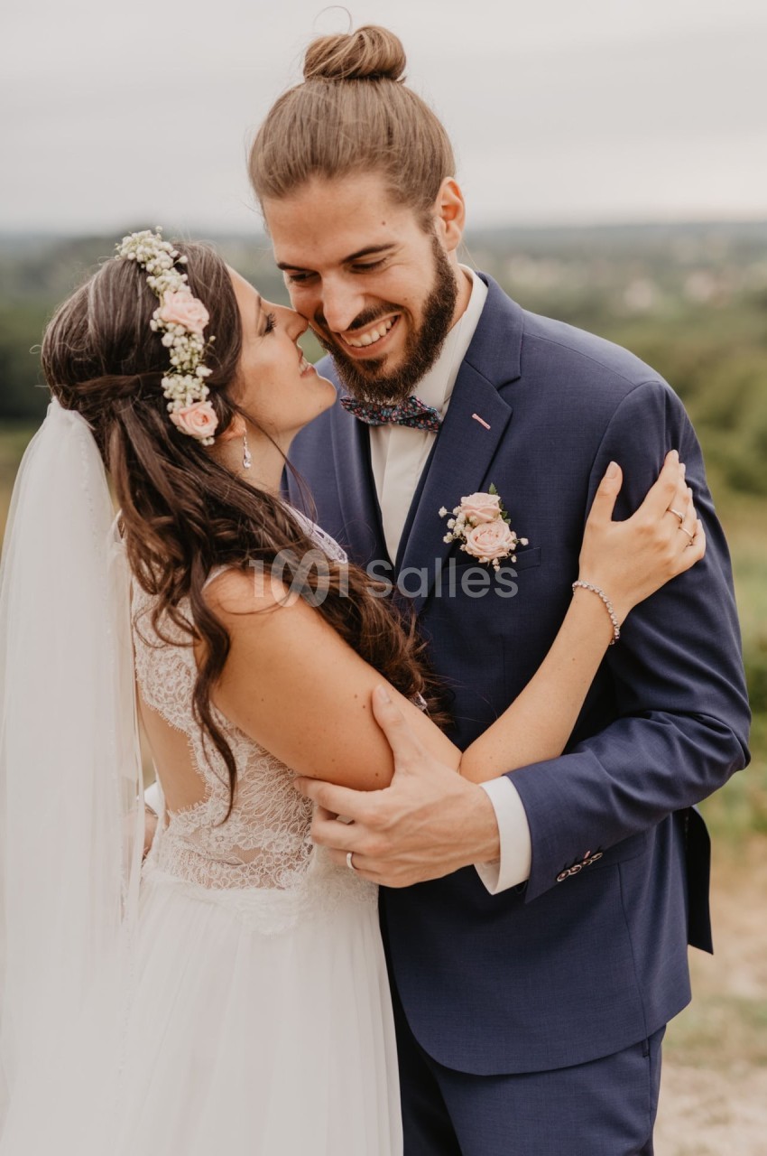 Un couple en tenue de mariage s'enlace tendrement en extérieur, avec un paysage naturel en arrière-plan.