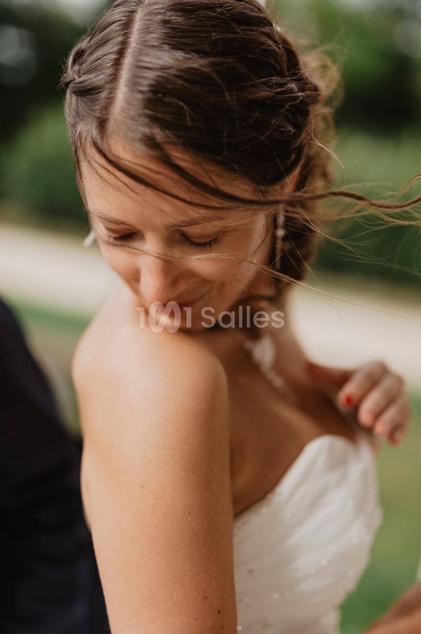 Femme en robe blanche regardant son épaule, cheveux légèrement décoiffés par le vent, arrière-plan flou verdoyant.