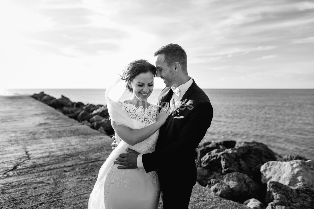 Un couple en tenue de mariage sourit et s'enlace près d'une jetée face à la mer, sous un ciel dégagé.