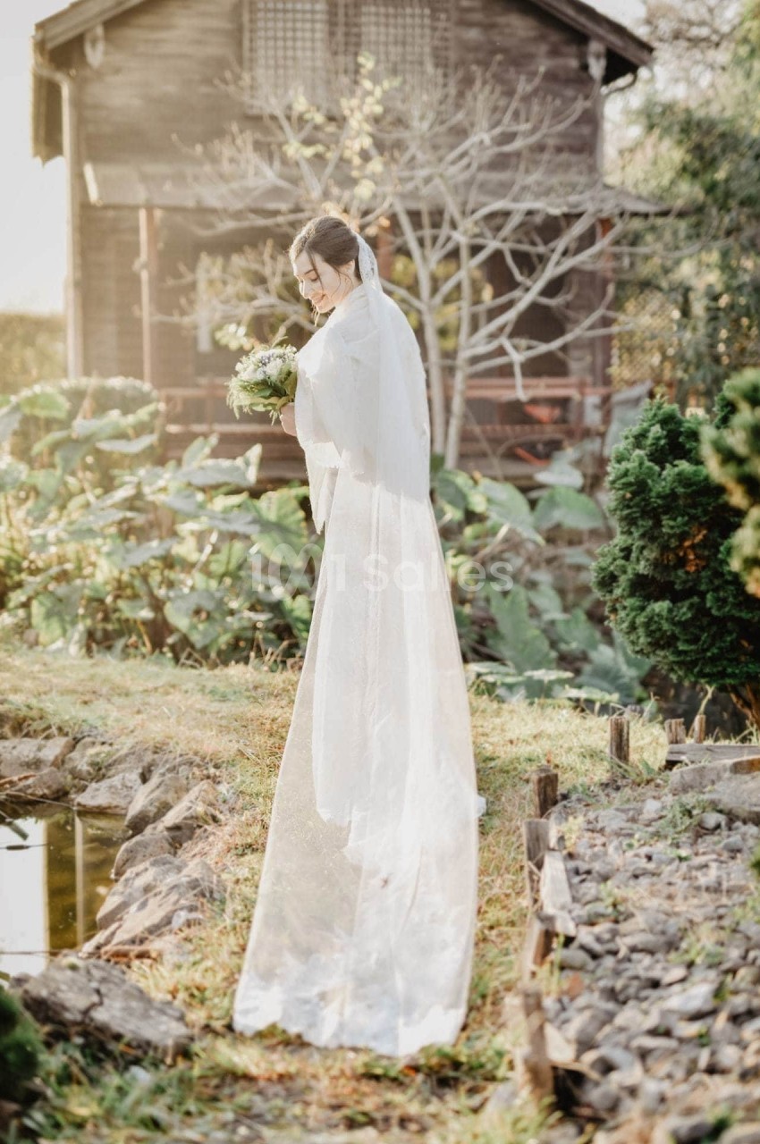 Une femme en robe de mariée blanche tient un bouquet près d'un étang, devant une maison en bois et un jardin verdoyant.