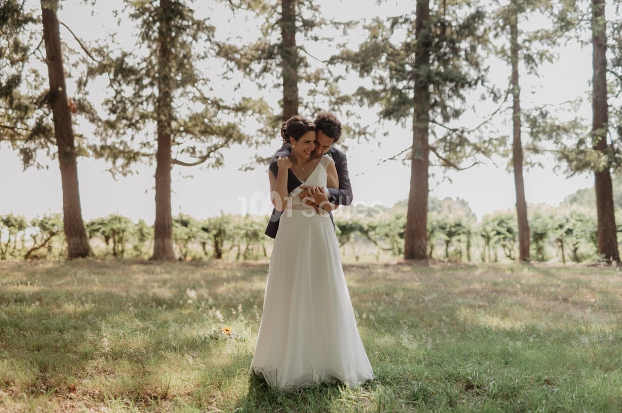 Un couple en tenue de mariage s'enlace dans un champ ensoleillé bordé d'arbres.