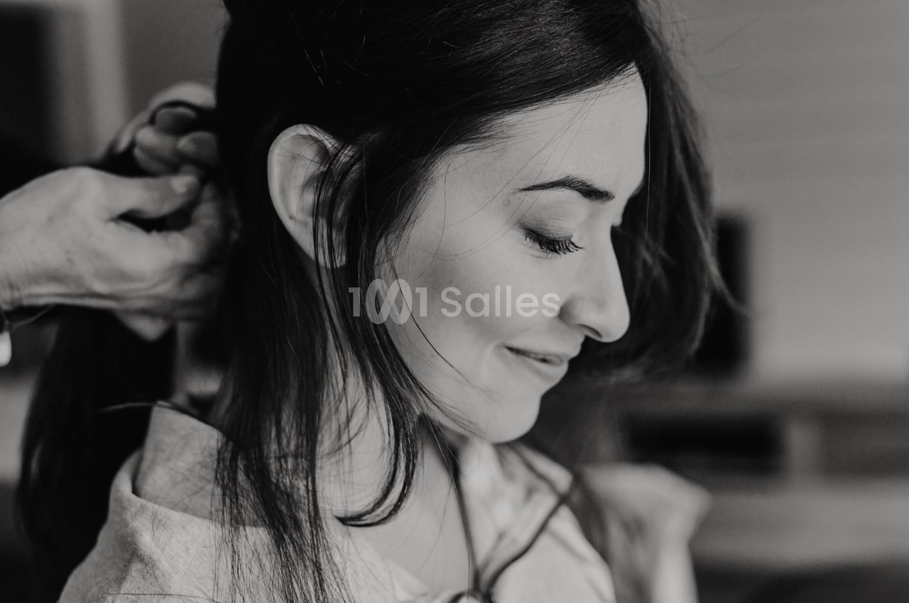 Femme souriante de profil, cheveux attachés par une autre personne dans un cadre intérieur.