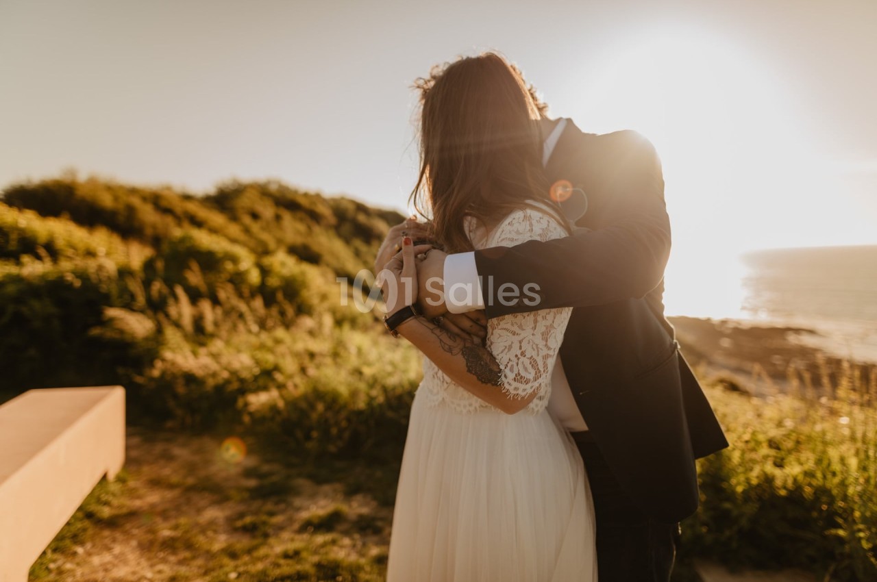 Un couple s'enlace tendrement au coucher du soleil, près d'un paysage naturel verdoyant et d'une étendue d'eau.