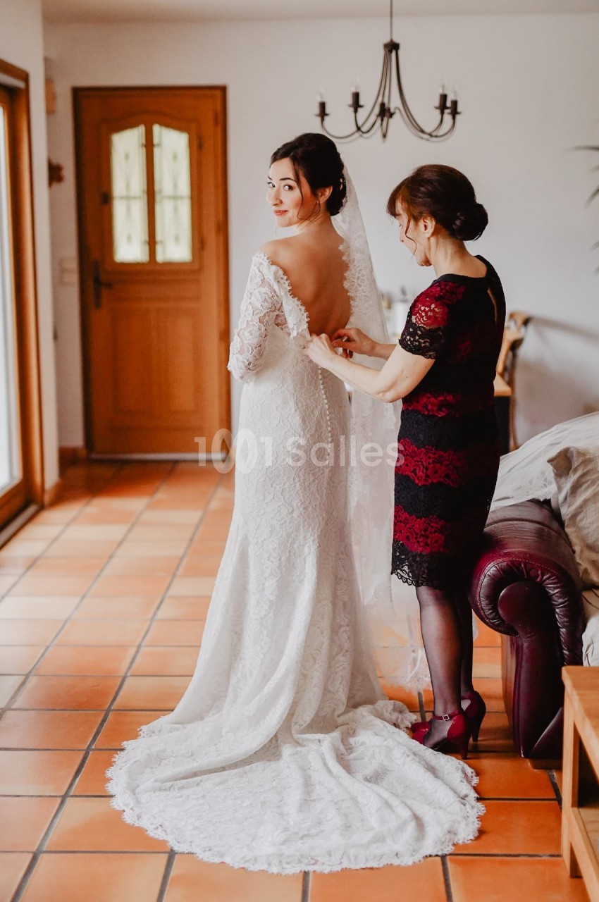 Une femme aide une mariée à ajuster sa robe blanche dans une pièce lumineuse avec un sol en carrelage.