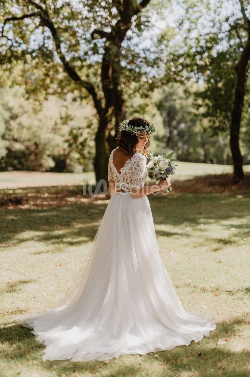 Une femme en robe de mariée blanche tient un bouquet, debout dans un parc ensoleillé entouré d'arbres.