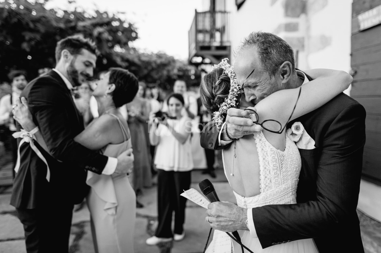 Un père ému enlace sa fille en robe de mariée, tandis qu'un couple danse en arrière-plan lors d'une réception.