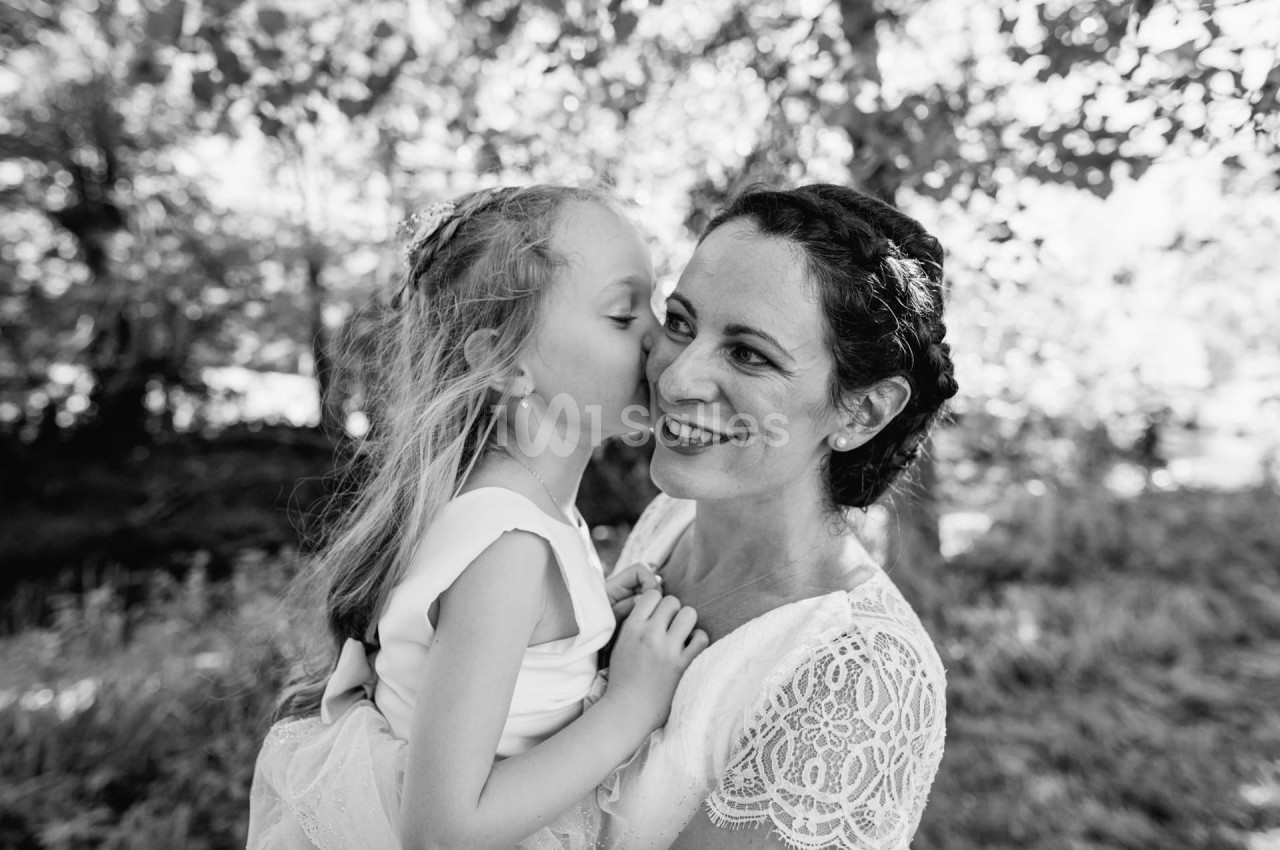Une femme souriante porte une fillette qui lui chuchote à l'oreille dans un cadre naturel.