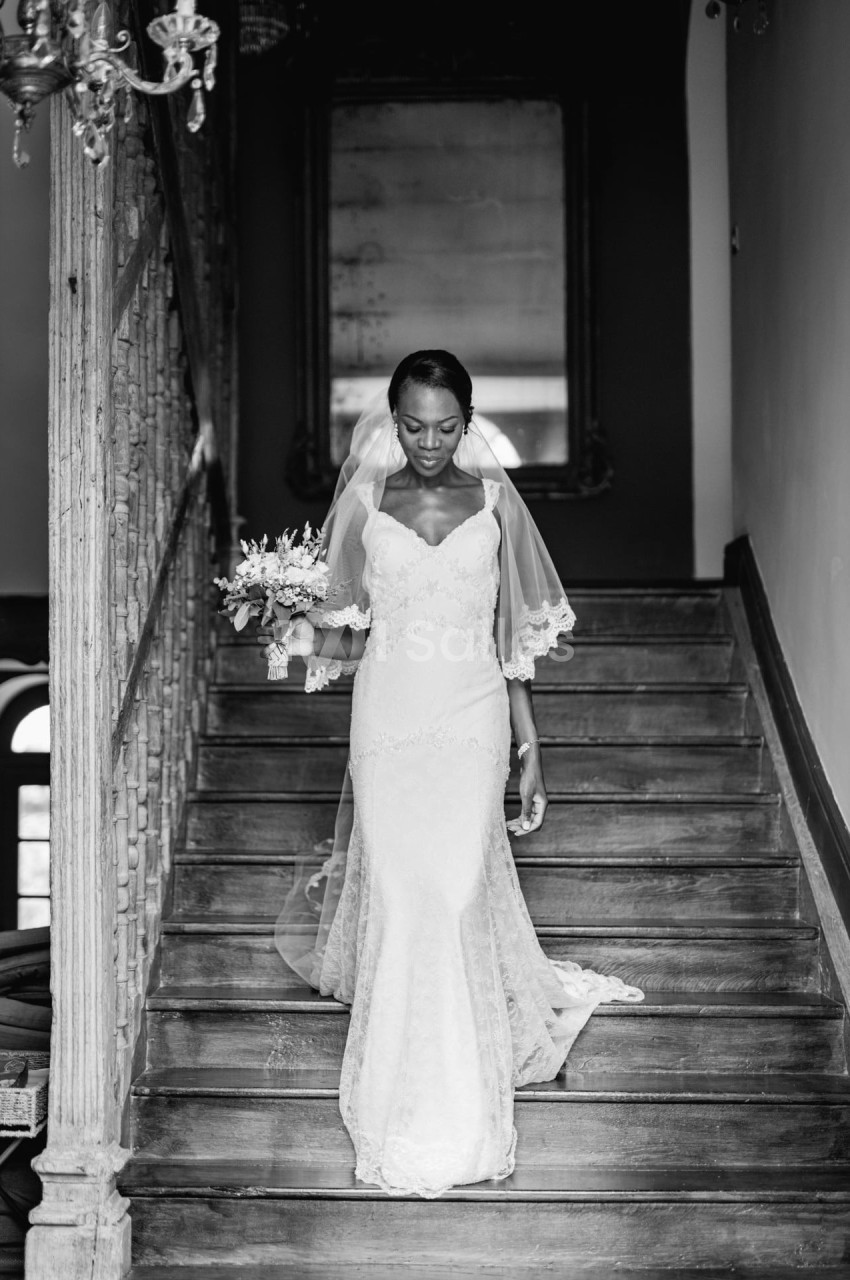 Une mariée en robe blanche et voile descend un escalier en bois, tenant un bouquet de fleurs.