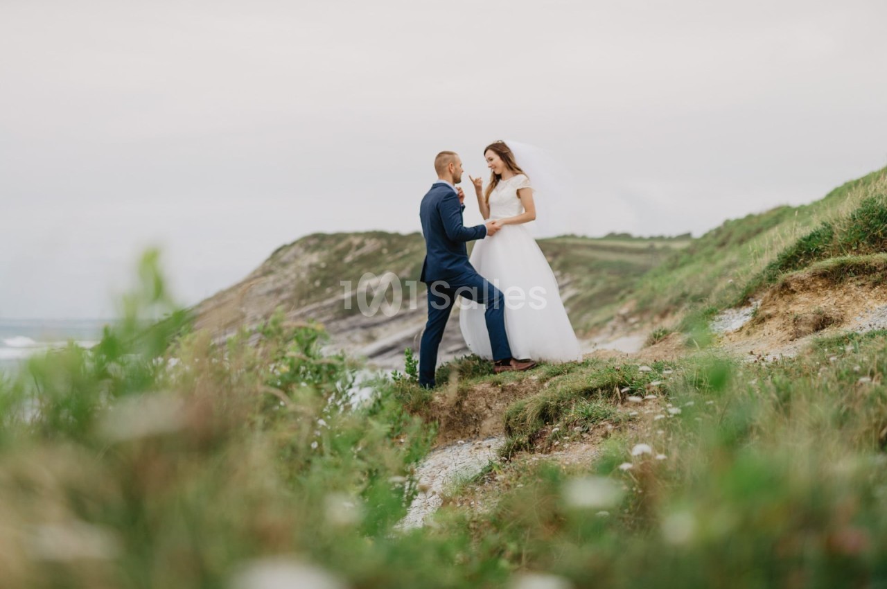 Un couple en tenue de mariage pose sur une colline verdoyante avec un paysage côtier en arrière-plan.