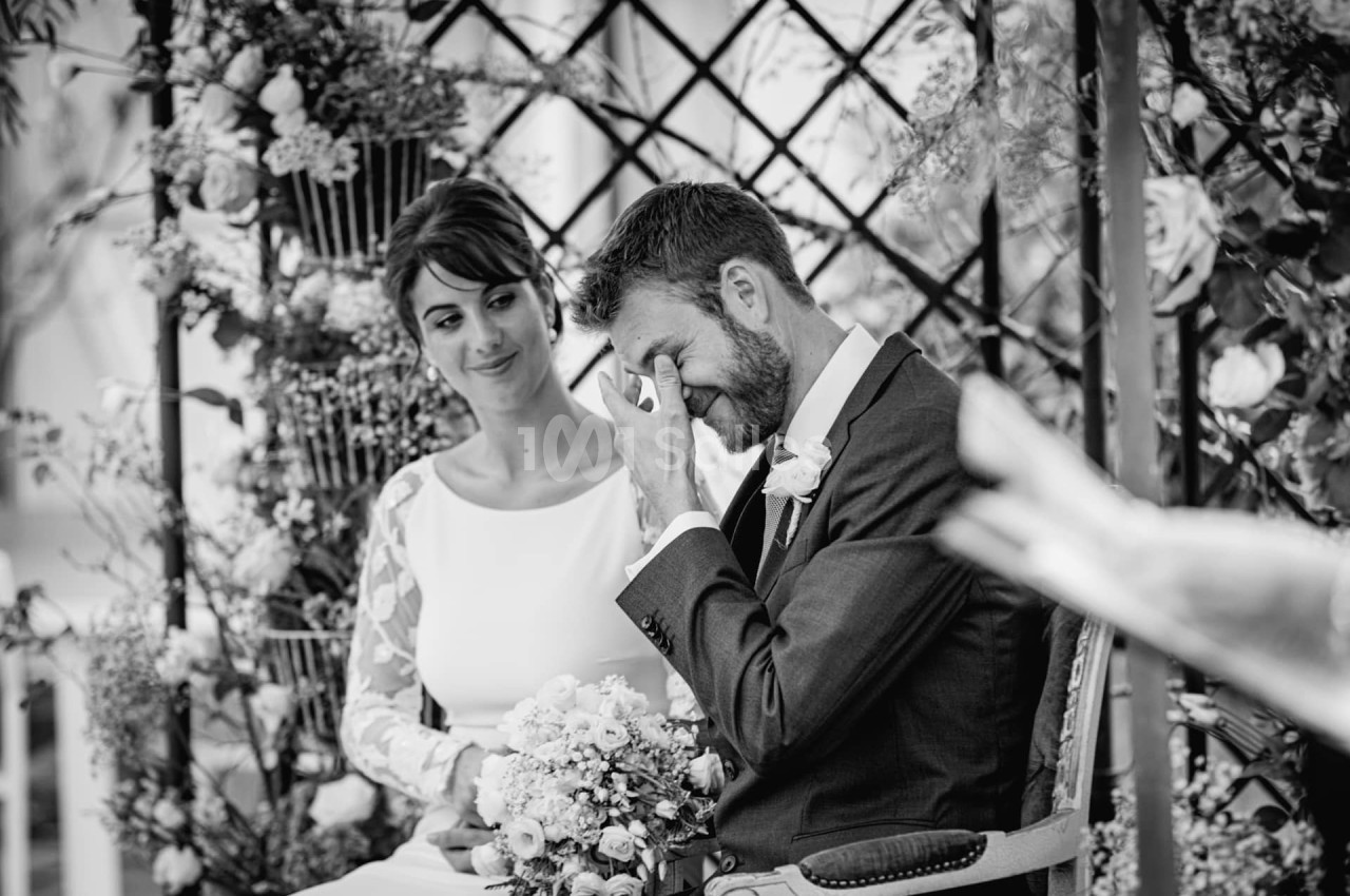 Un couple assis lors d'une cérémonie, l'homme ému essuie une larme tandis que la femme le regarde avec tendresse.