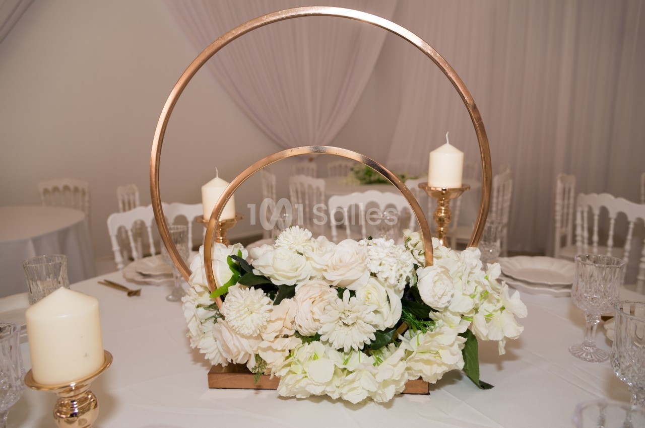 Centre de table avec des fleurs blanches, des bougies et des cercles dorés sur une nappe blanche.