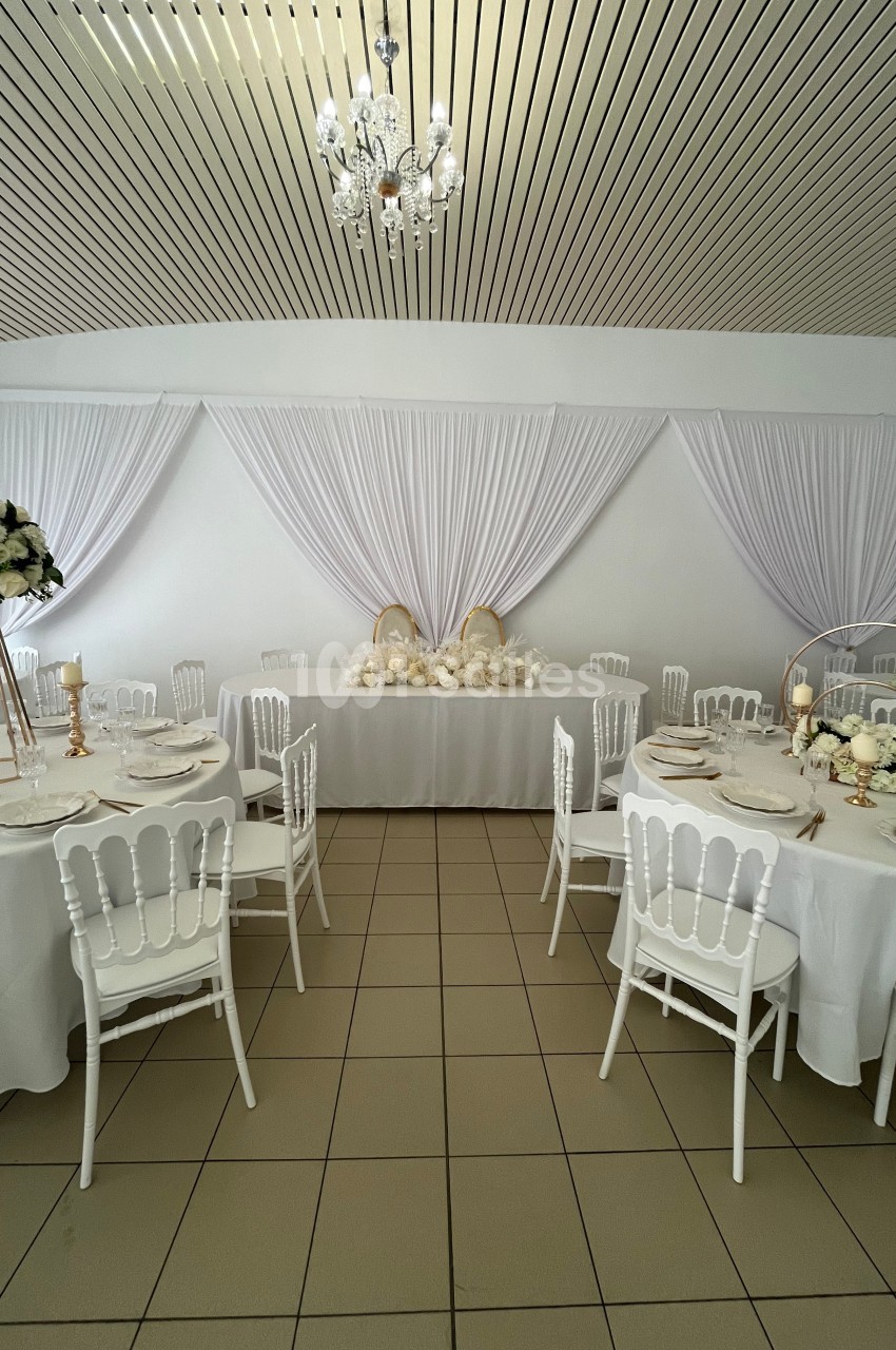 Salle de réception décorée en blanc avec des tables rondes, des chaises élégantes et des arrangements floraux.