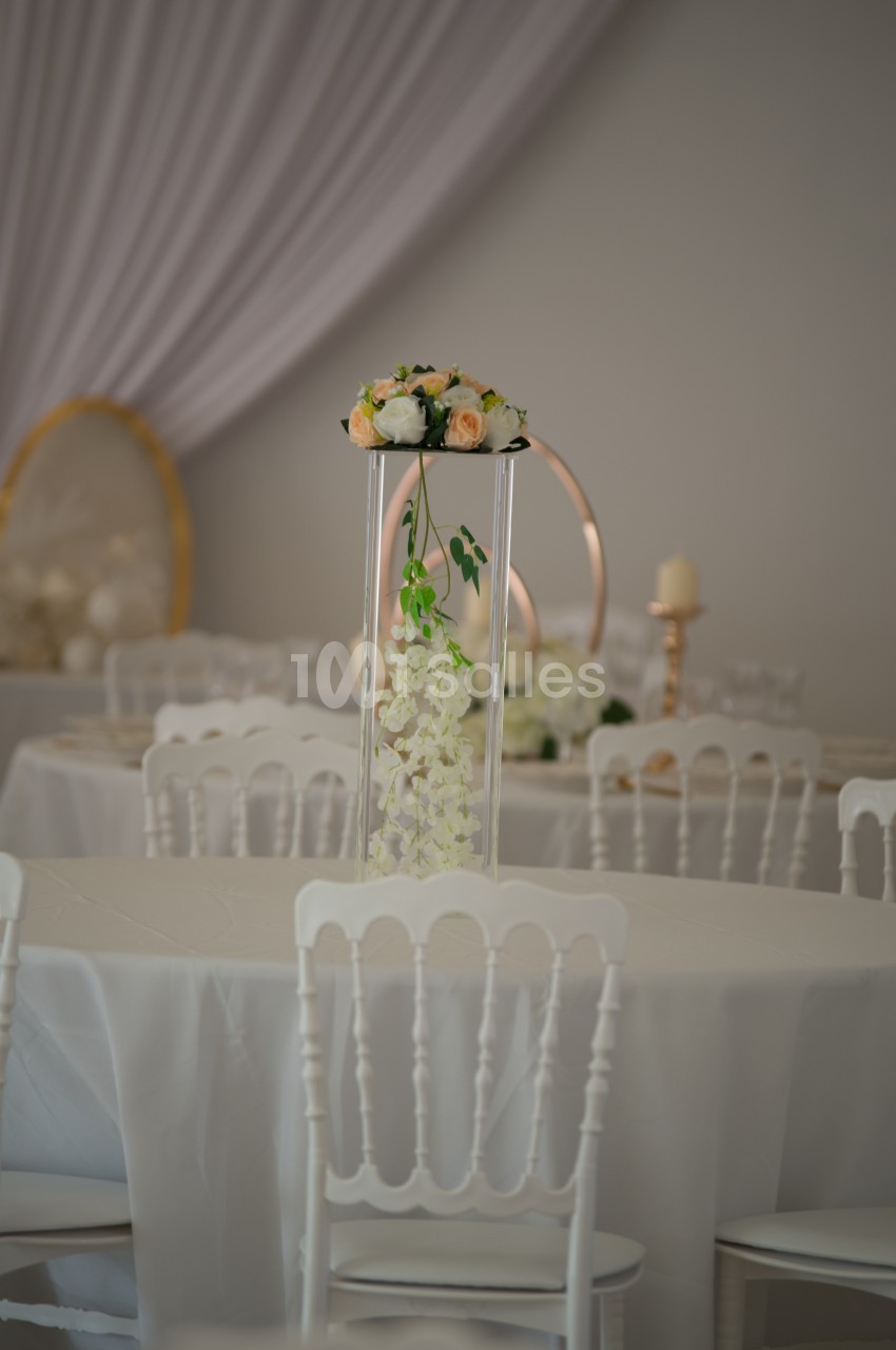 Centre de table floral avec roses et orchidées blanches, posé sur un support transparent dans une salle décorée.