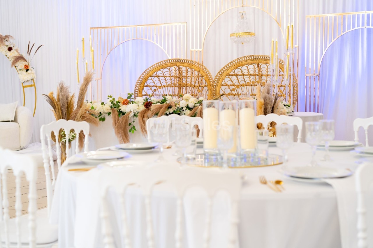Salle de réception décorée avec des chaises blanches, une table dressée et un fond orné de fleurs et d'éléments dorés.