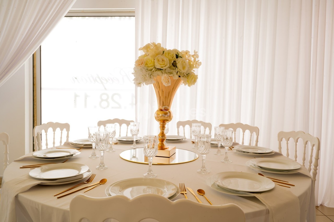 Table ronde élégamment dressée avec une nappe blanche, vaisselle raffinée et un vase doré rempli de fleurs blanches.