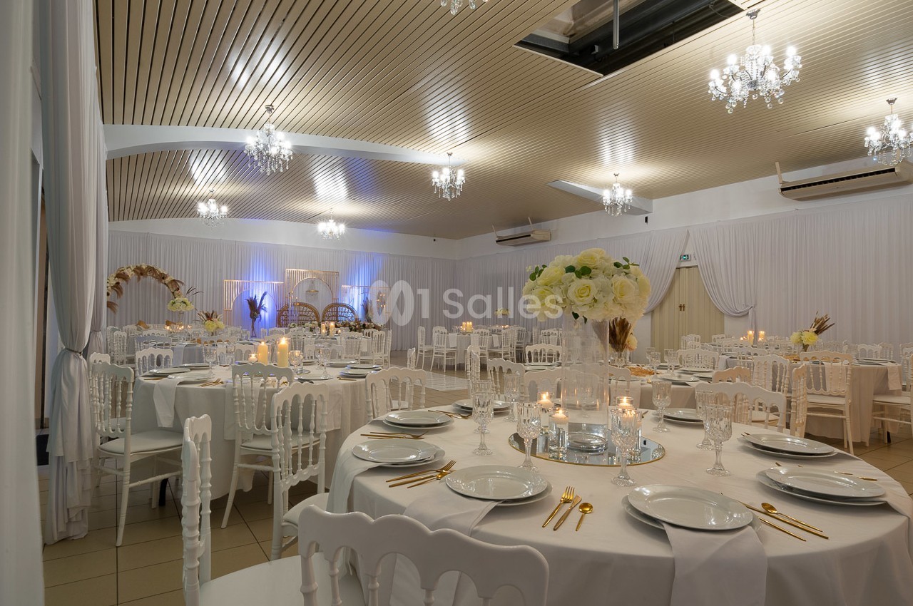 Salle de réception élégamment décorée avec des tables rondes, nappes blanches, chandeliers et éclairage chaleureux.