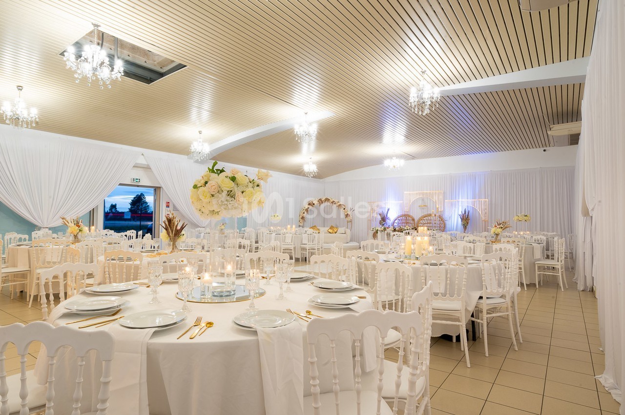 Salle de réception élégamment décorée avec des tables rondes, des chaises blanches et des arrangements floraux.