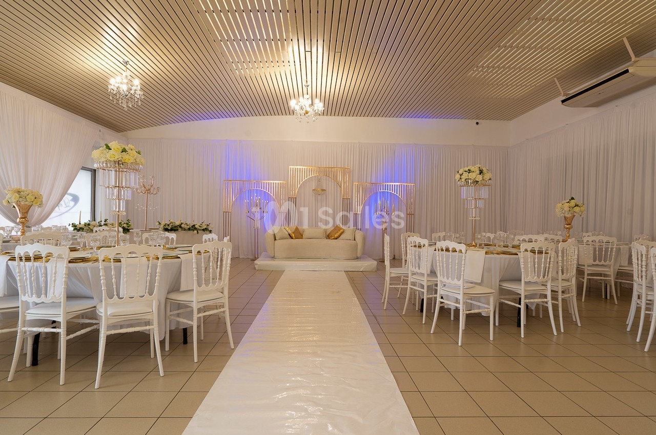 Salle de réception décorée avec des tables rondes, des chaises blanches et une estrade ornée de fleurs et de lumière.
