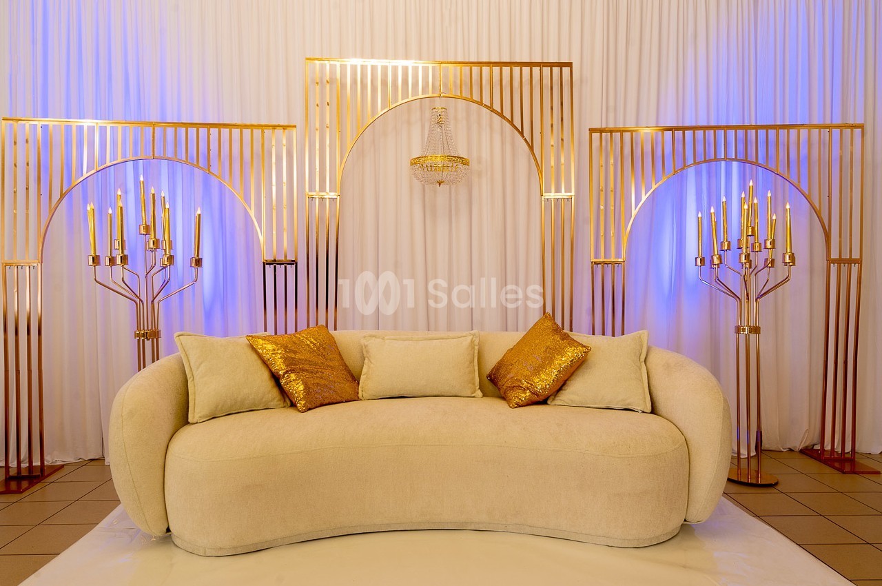Canapé beige avec coussins dorés, entouré de chandeliers et d'arcs métalliques dorés sur fond de rideaux blancs.
