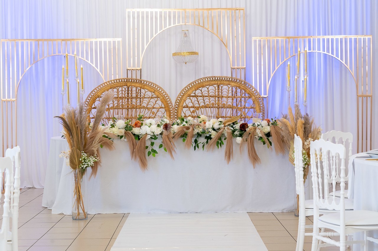 Décoration de table de mariage avec chaises en rotin, fleurs blanches et pampas, devant un fond blanc et doré.