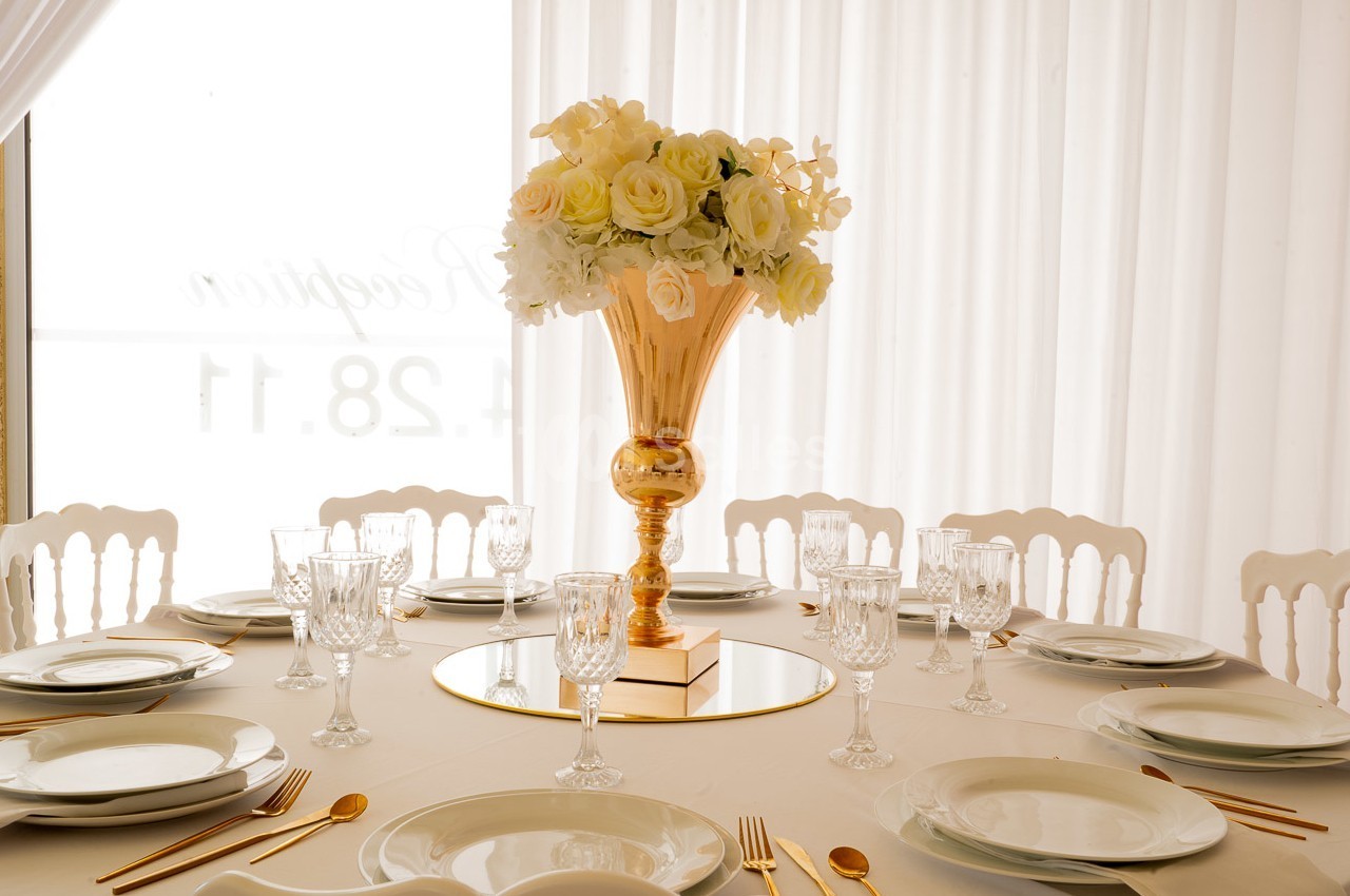 Table élégamment dressée avec vaisselle blanche, couverts dorés et un centre de table floral dans un vase doré.