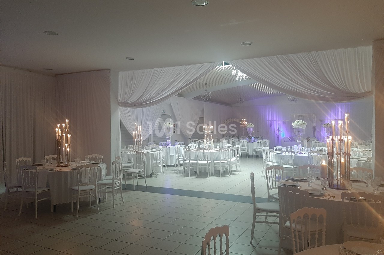 Salle de réception décorée avec des tables rondes, des chaises blanches, des chandeliers et des drapés blancs au plafond.