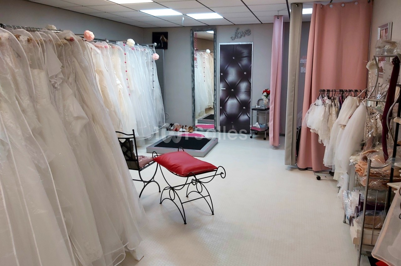 Salle d'essayage avec robes de mariée suspendues, miroir, fauteuil rouge et rideaux roses.