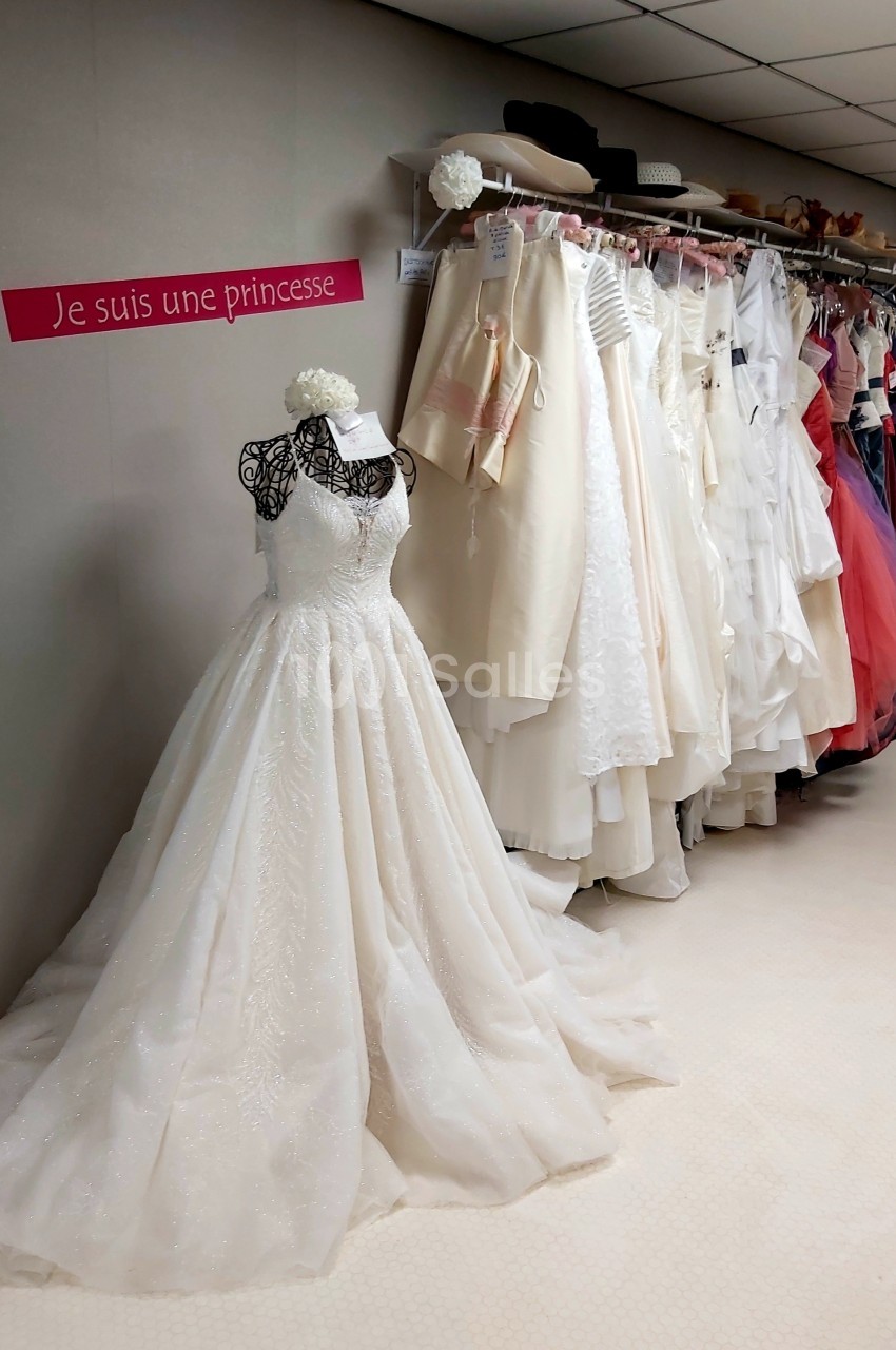Robe de mariée blanche exposée dans une boutique, entourée d'autres tenues suspendues sur des portants.