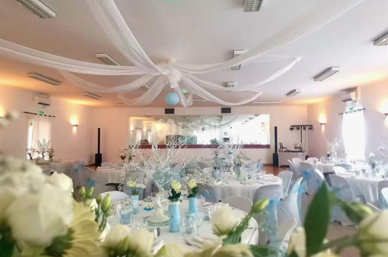 Salle de réception décorée en blanc et bleu avec tables dressées, fleurs et voilages suspendus au plafond.