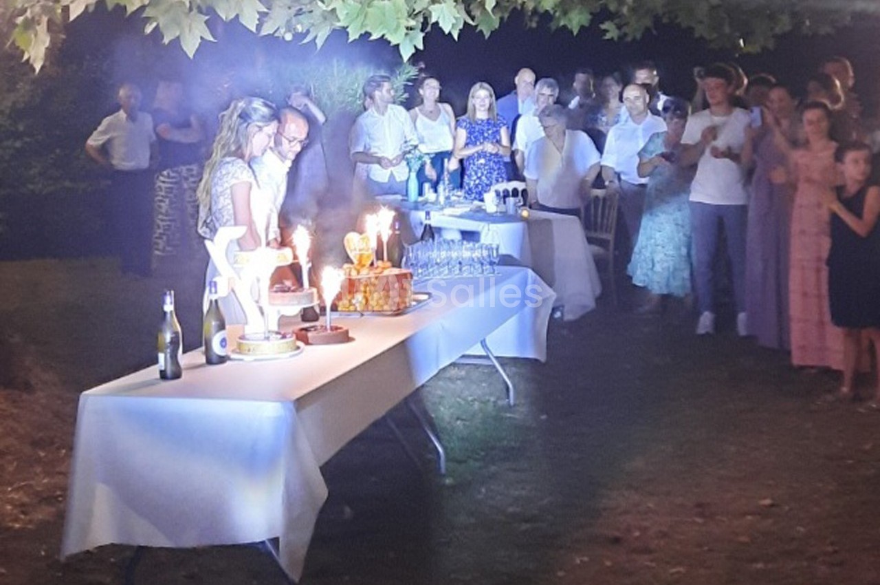 Des personnes rassemblées en extérieur autour de tables décorées, avec des gâteaux d'anniversaire éclairés par des bougies.