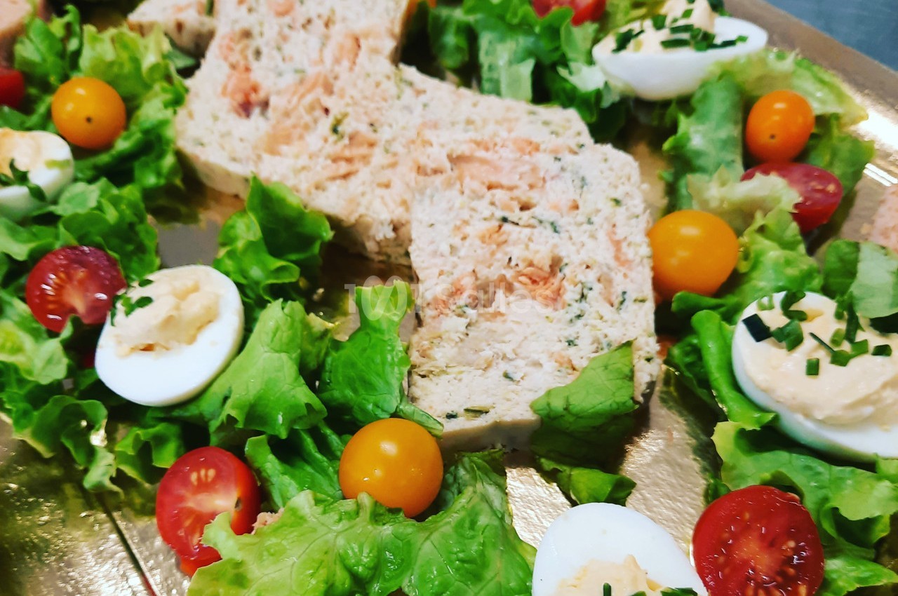 Terrine de poisson entourée de feuilles de salade, tomates cerises, œufs durs garnis et ciboulette sur un plateau doré.