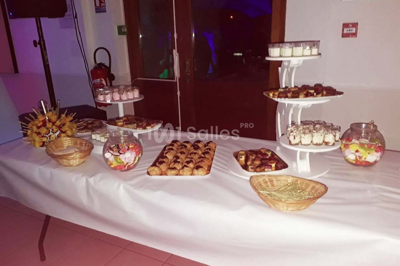 Table garnie de desserts variés, incluant verrines, pâtisseries, brochettes de fruits et bonbons, dans une salle éclairée.
