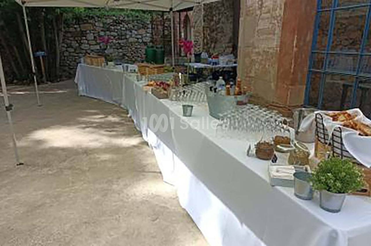 Tables dressées en extérieur avec nappes blanches, contenant verres, paniers de pain et divers éléments pour un buffet.