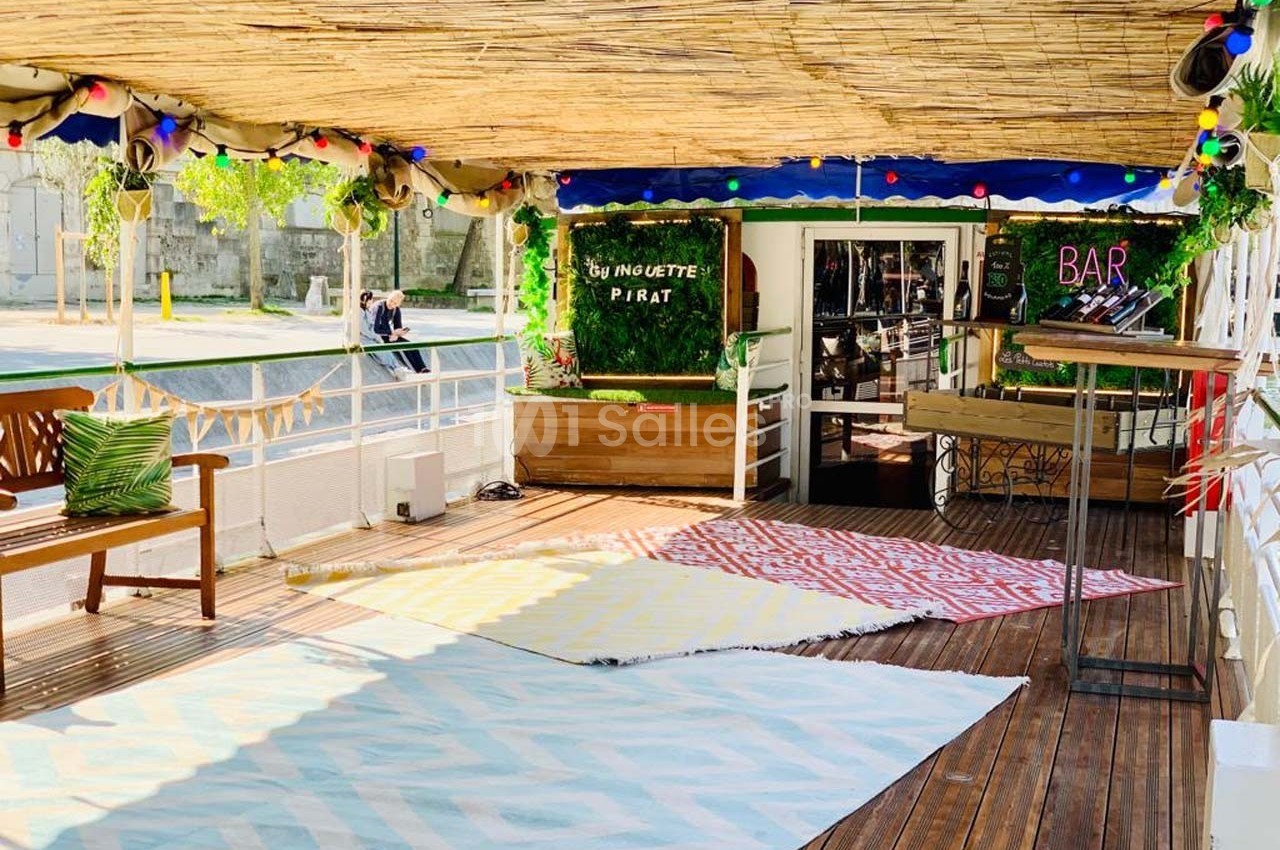 Terrasse en bois décorée avec des tapis colorés, des plantes et un bar en arrière-plan sous un toit en bambou.