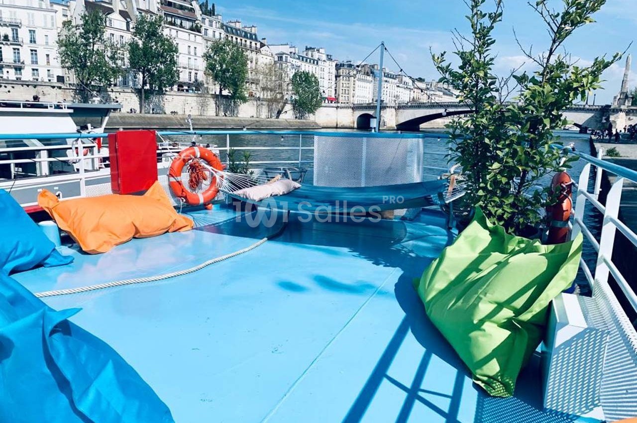 Pont d'un bateau avec coussins colorés, plantes en pot, bouée de sauvetage et vue sur des bâtiments en bord de rivière.