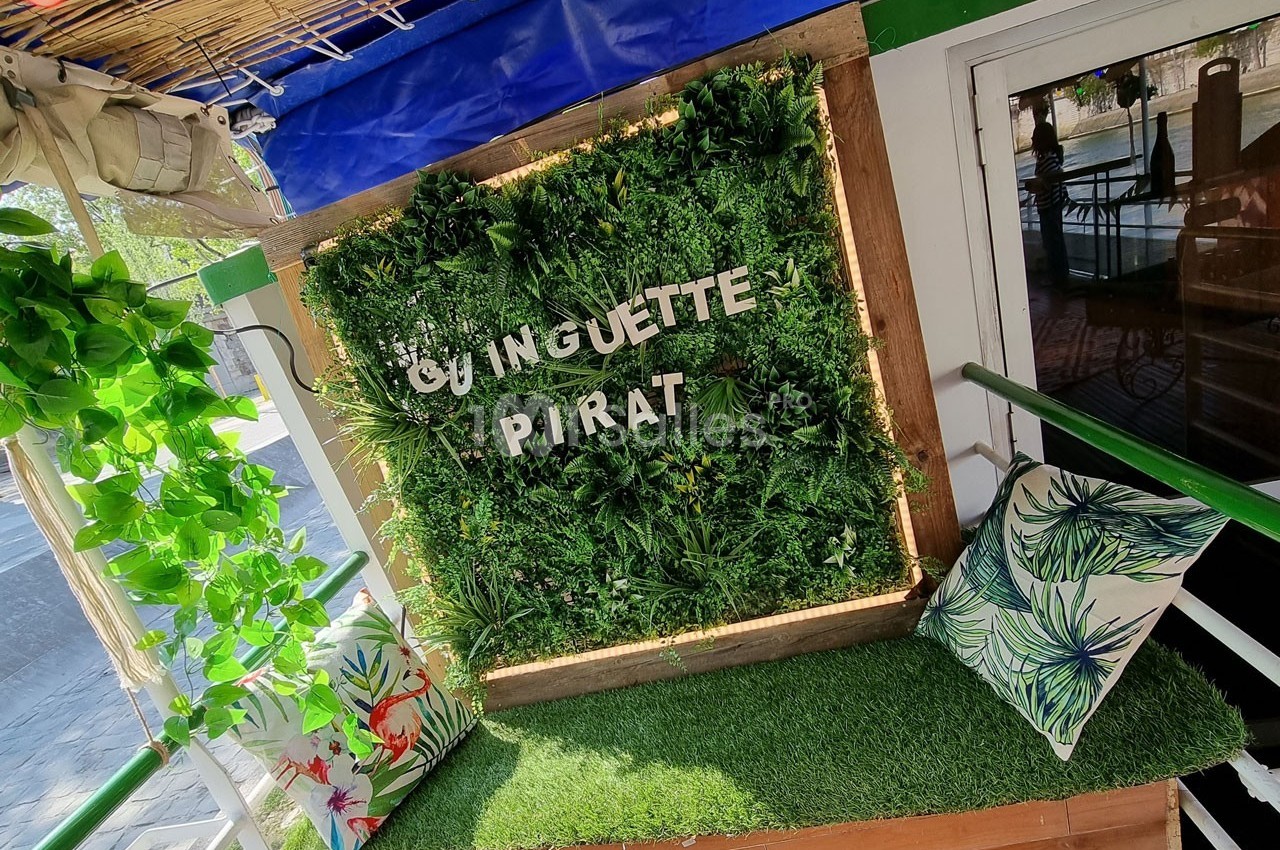 Mur végétal décoratif avec l'inscription ’Guinguette Pirate’, entouré de coussins à motifs tropicaux sur un banc.