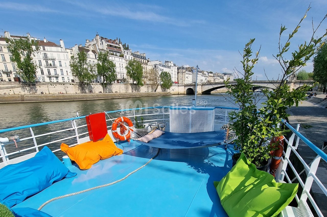 Terrasse d'un bateau avec coussins colorés, vue sur la Seine, un pont et des bâtiments parisiens en arrière-plan.