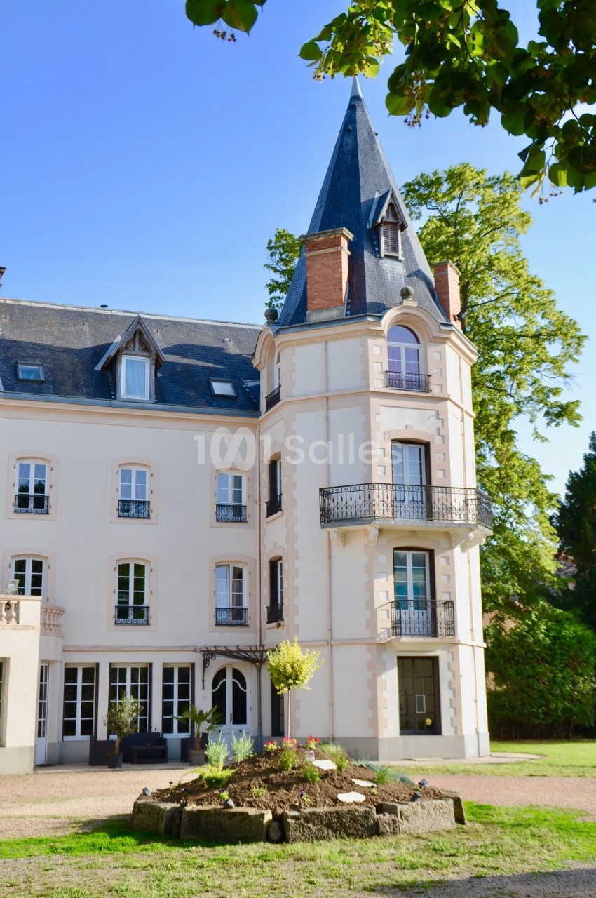 Façade d'un bâtiment ancien avec une tour, des balcons et des fenêtres, entouré de végétation et d'un parterre fleuri.