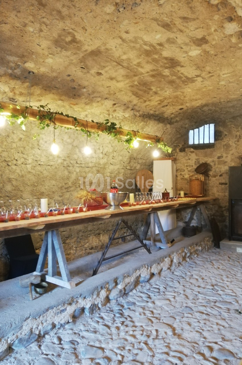 Table en bois avec verres et bouteilles alignés dans une cave voûtée en pierre, éclairée par des guirlandes lumineuses.