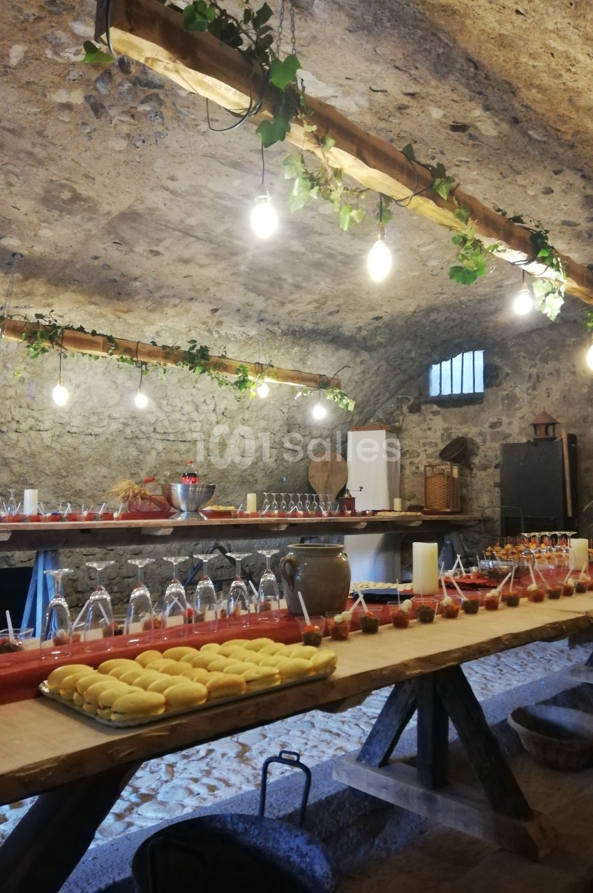 Salle voûtée en pierre avec une longue table en bois garnie de verrines, plats et décorations lumineuses.