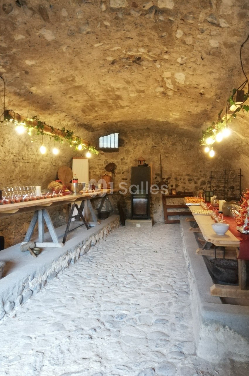 Salle voûtée en pierre aménagée avec des tables garnies de plats, boissons et décorations lumineuses.