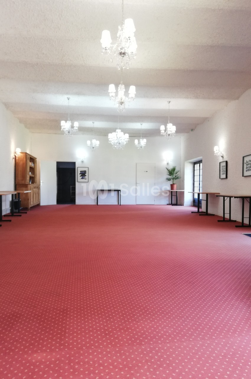 Salle spacieuse avec moquette rouge, murs blancs, lustres suspendus et tables alignées le long des murs.