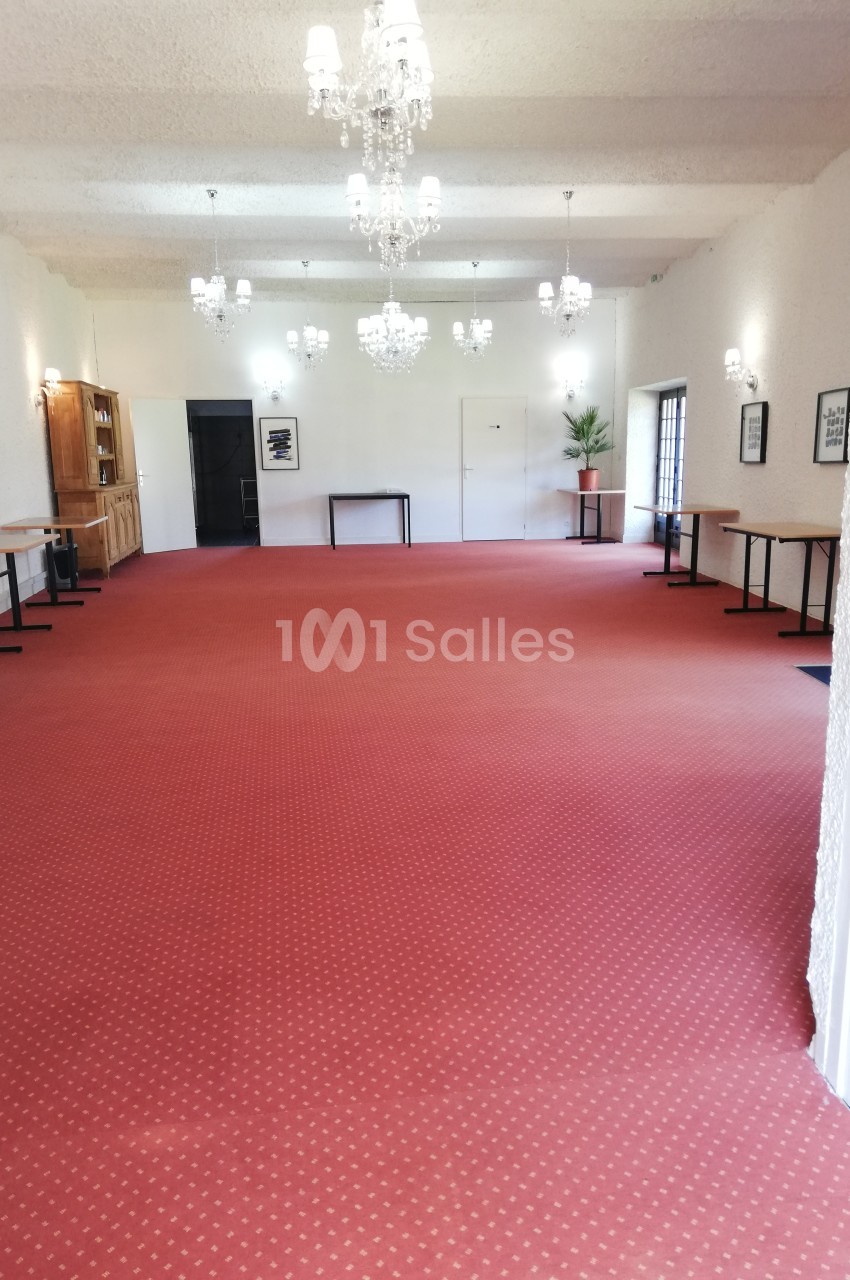 Salle spacieuse avec moquette rouge, murs blancs, lustres suspendus et tables alignées le long des côtés.