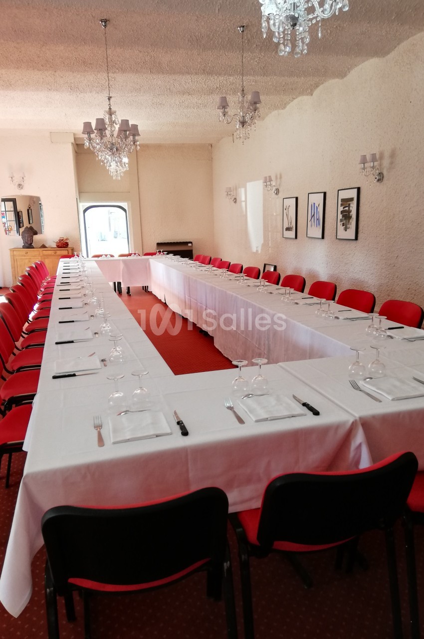 Salle de réunion avec tables en U couvertes de nappes blanches, chaises rouges et verres disposés pour un événement.