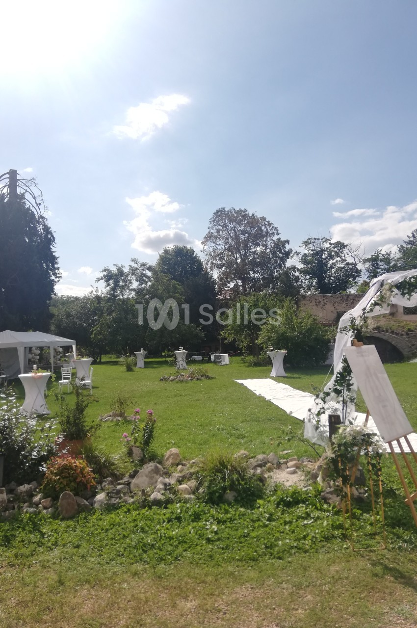 Jardin ensoleillé aménagé pour un événement avec tables hautes, tente blanche et allée décorée de fleurs.
