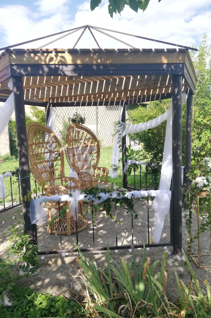 Tonnelle décorée de voilages blancs et de fleurs, avec des fauteuils en rotin, dans un jardin ensoleillé.