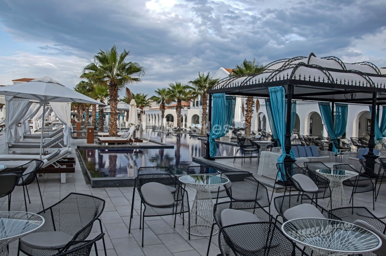 Terrasse d'un hôtel avec piscine, chaises, tables, lits balinais, palmiers et ciel nuageux en arrière-plan.