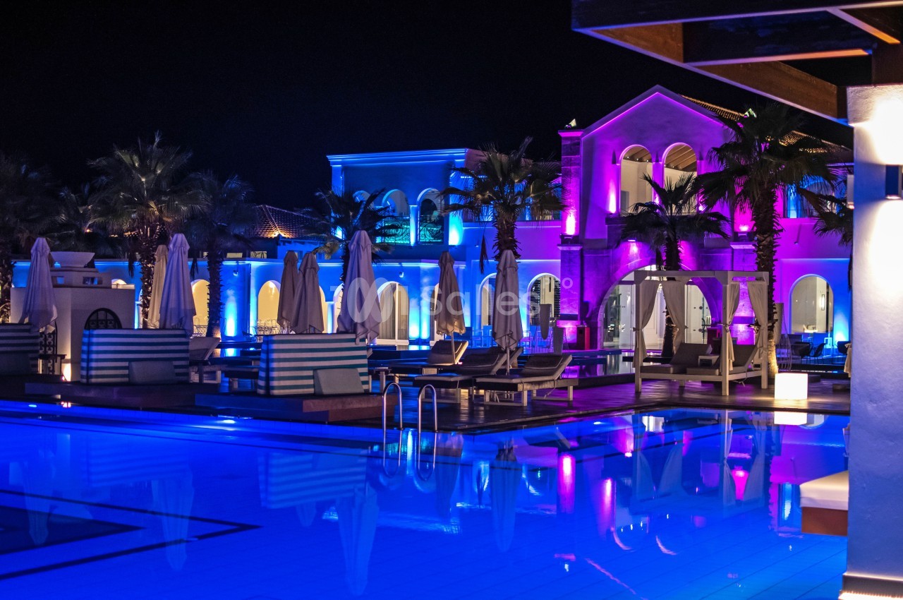 Piscine éclairée de nuit entourée de transats, parasols fermés et bâtiments illuminés aux tons colorés.