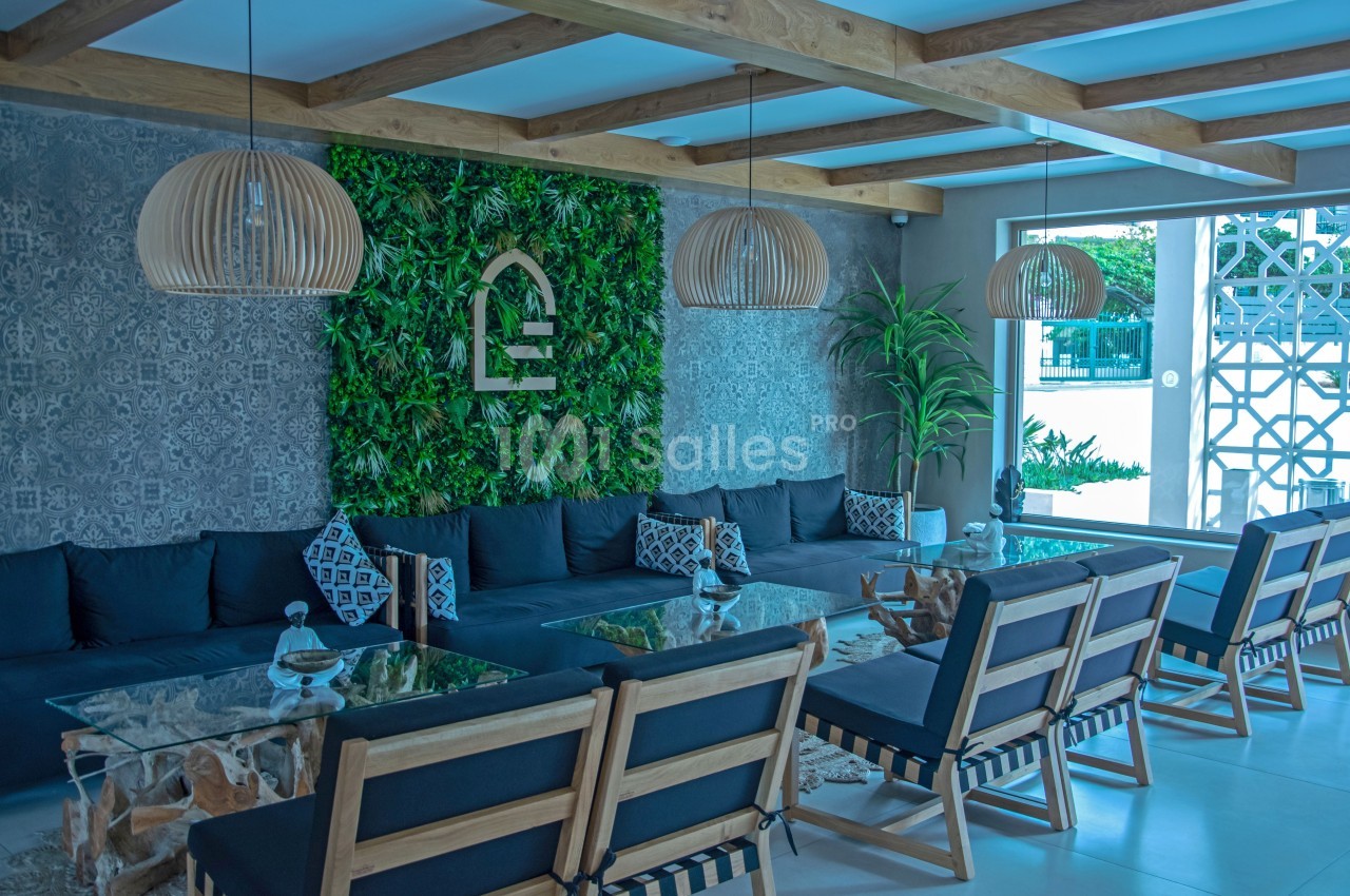 Salon moderne avec murs végétalisés, canapés noirs, tables en verre et chaises en bois sous des luminaires suspendus.